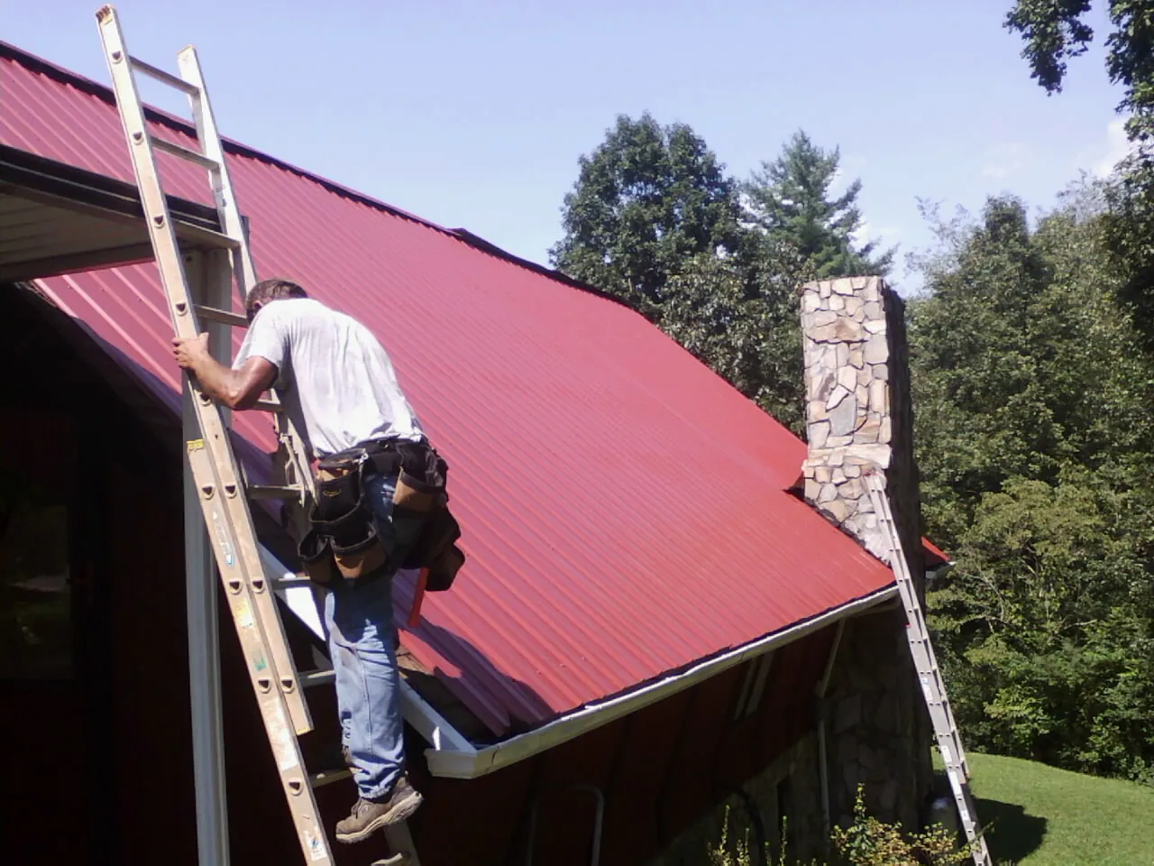 Roofer on a ladder beside a red metal roof and stone chimney — Soffit & Fascia Repair by Roofing Brothers in Lake Elsinore